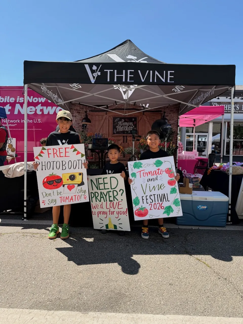 Three children holding the free photo booth, prayer, and Tomato & Vine Festival signs in front of the booth