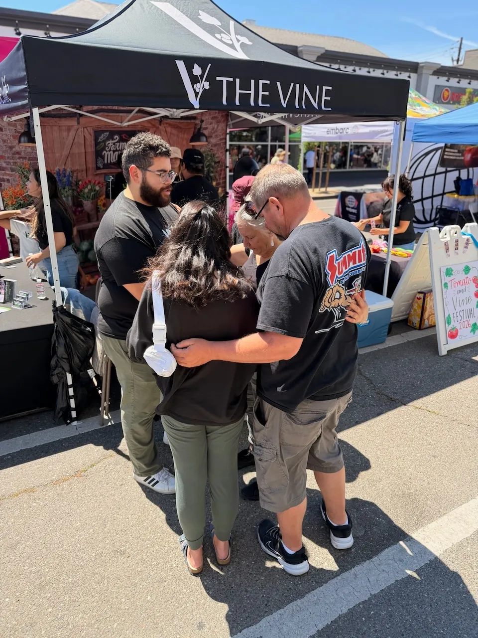 A group praying together, arms around one another, under The Vine's canopy at the Tomato & Vine Festival